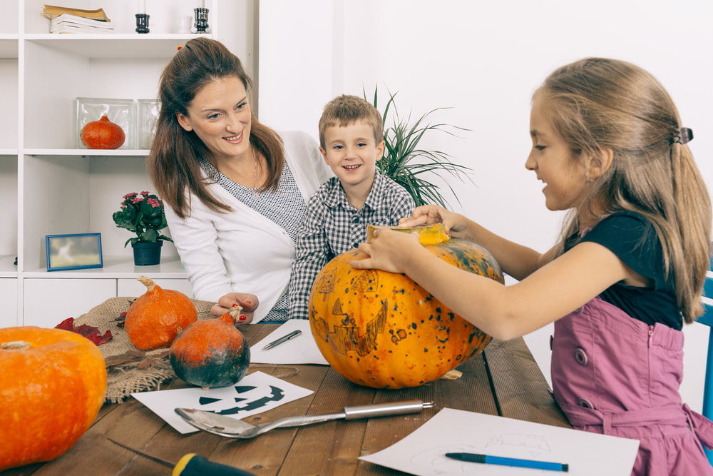 Lady, little boy and little girl are standing around brown table with different size pumpkins on the table, the little girl is holding the pumpkin and taking the top off the pumpkin